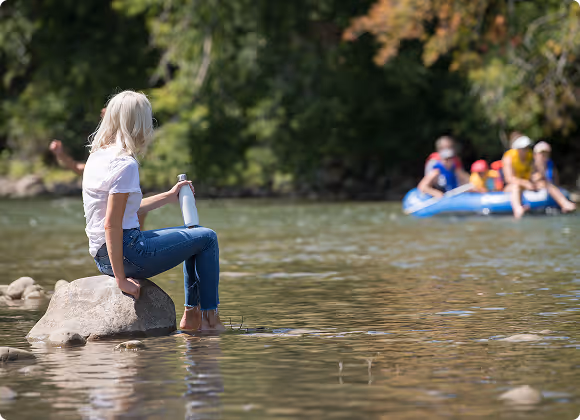 Woman with blonde hair sitting barefoot on a rock in a shallow river holding a water bottle, with a group of people rafting in the background.