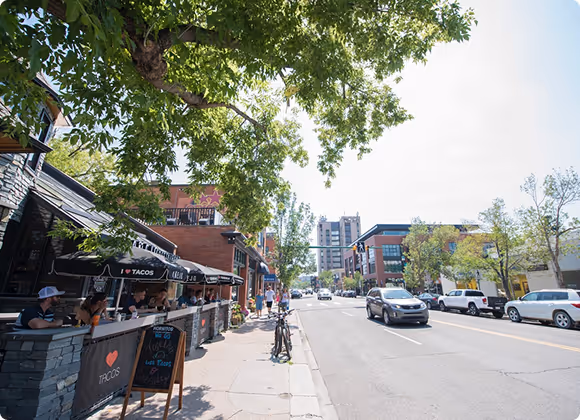 Street scene with outdoor seating under black awnings for a taco restaurant, people dining, parked cars, and buildings under a large leafy tree.