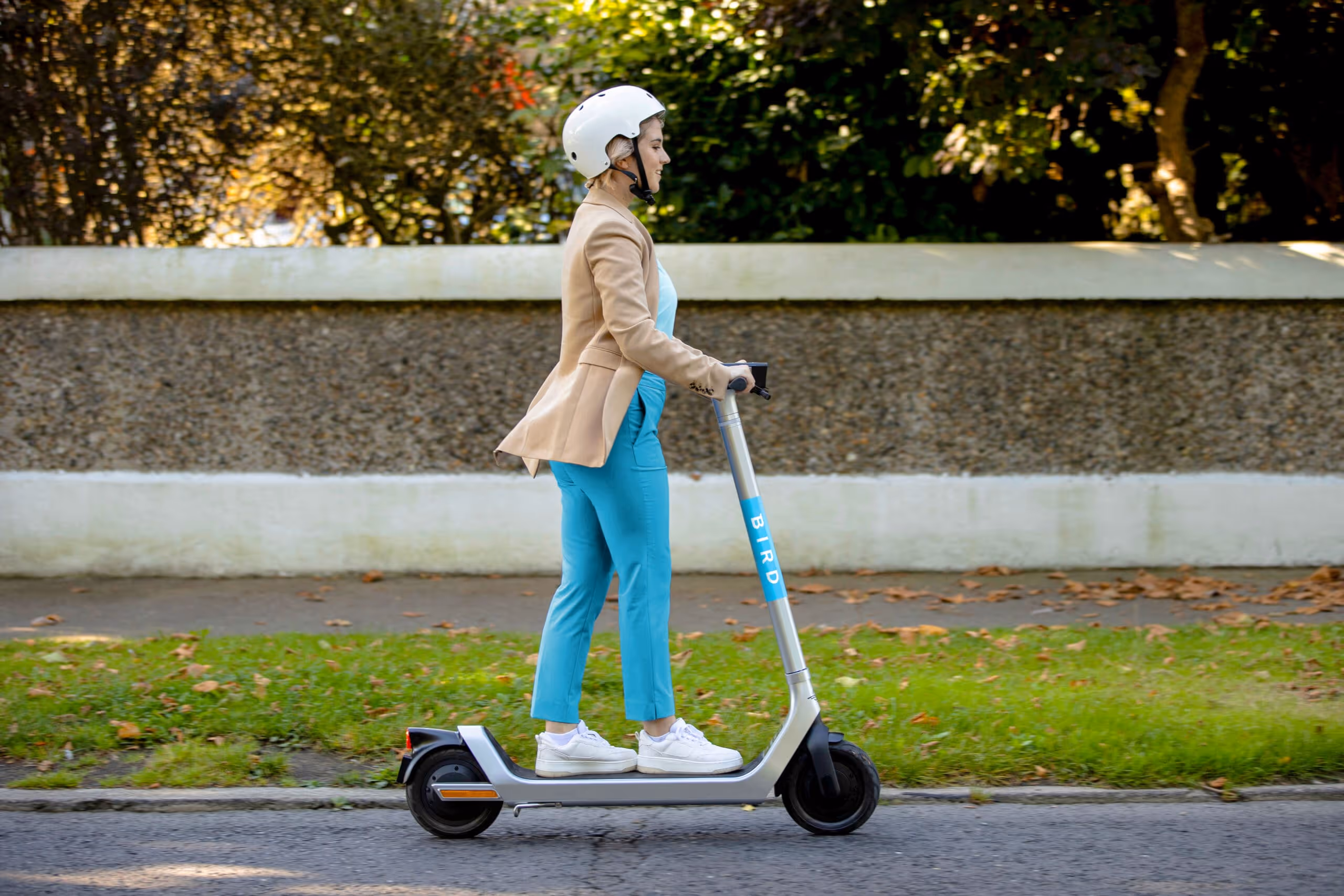 Woman wearing a helmet riding a Bird electric scooter on a paved road beside a grassy area and a stone wall.