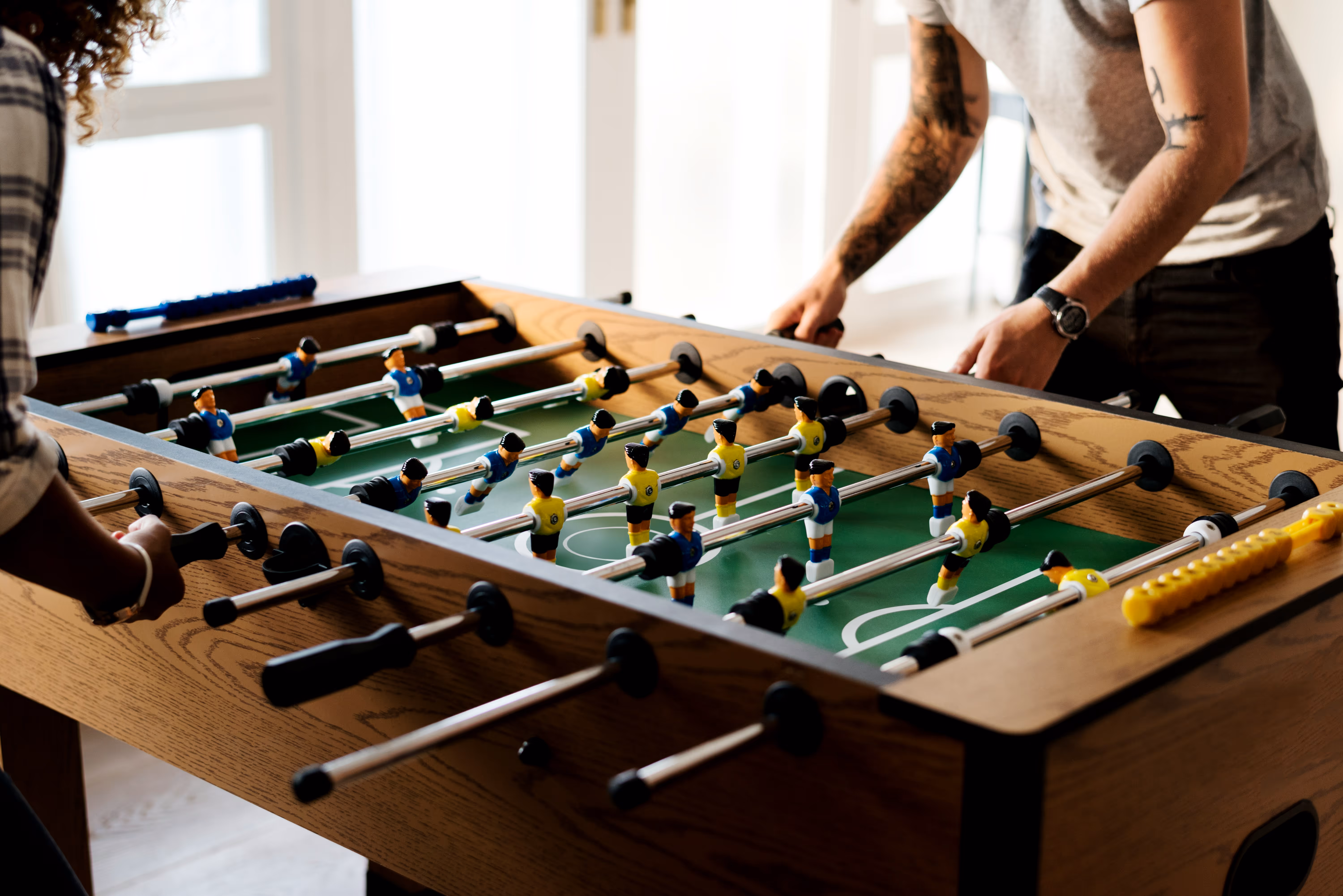 Two people playing a foosball game on a wooden table with yellow and blue player figures in a bright room.