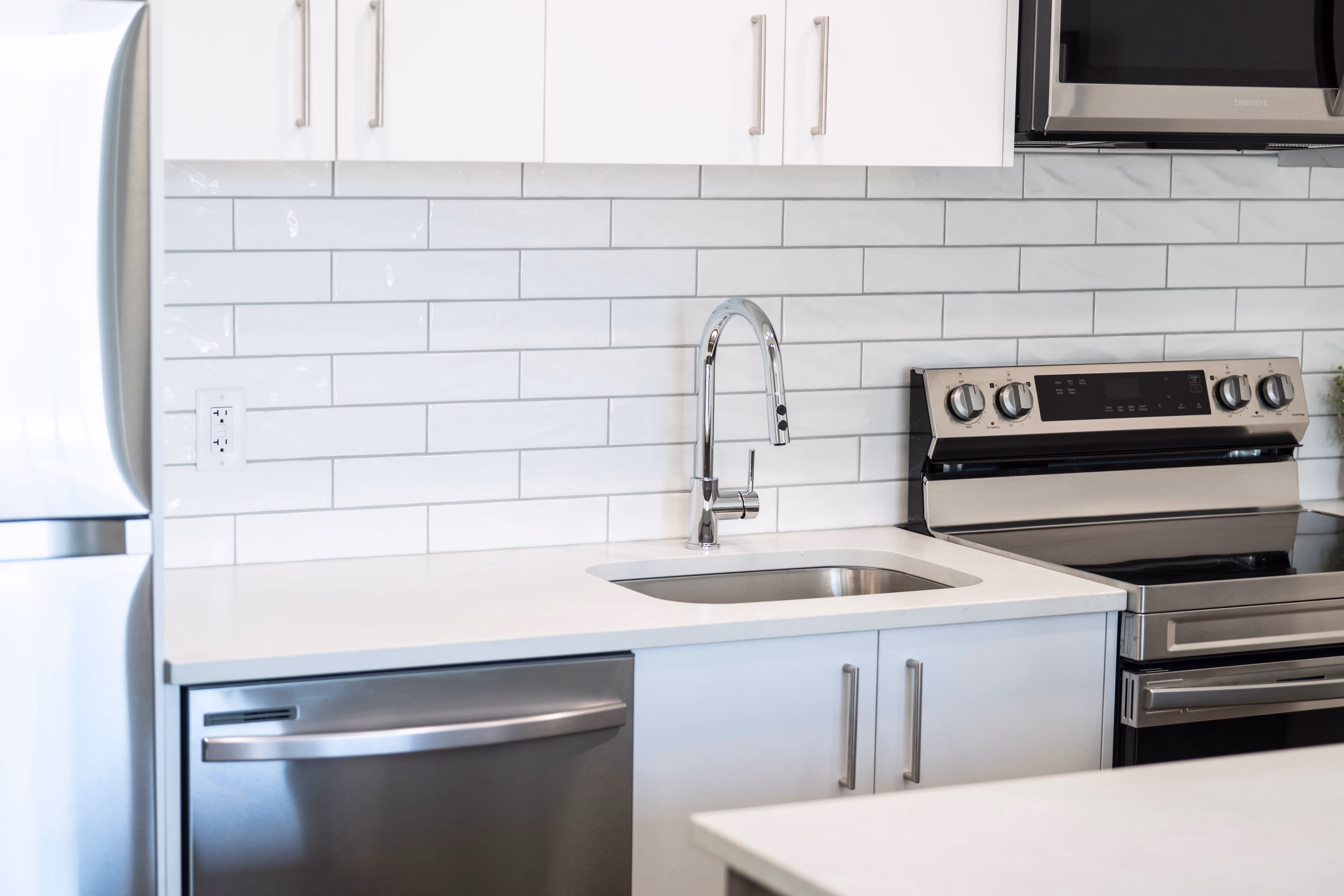 Modern kitchen with stainless steel refrigerator, built-in microwave, stove, sink, gray and white cabinets, and white tiled backsplash.