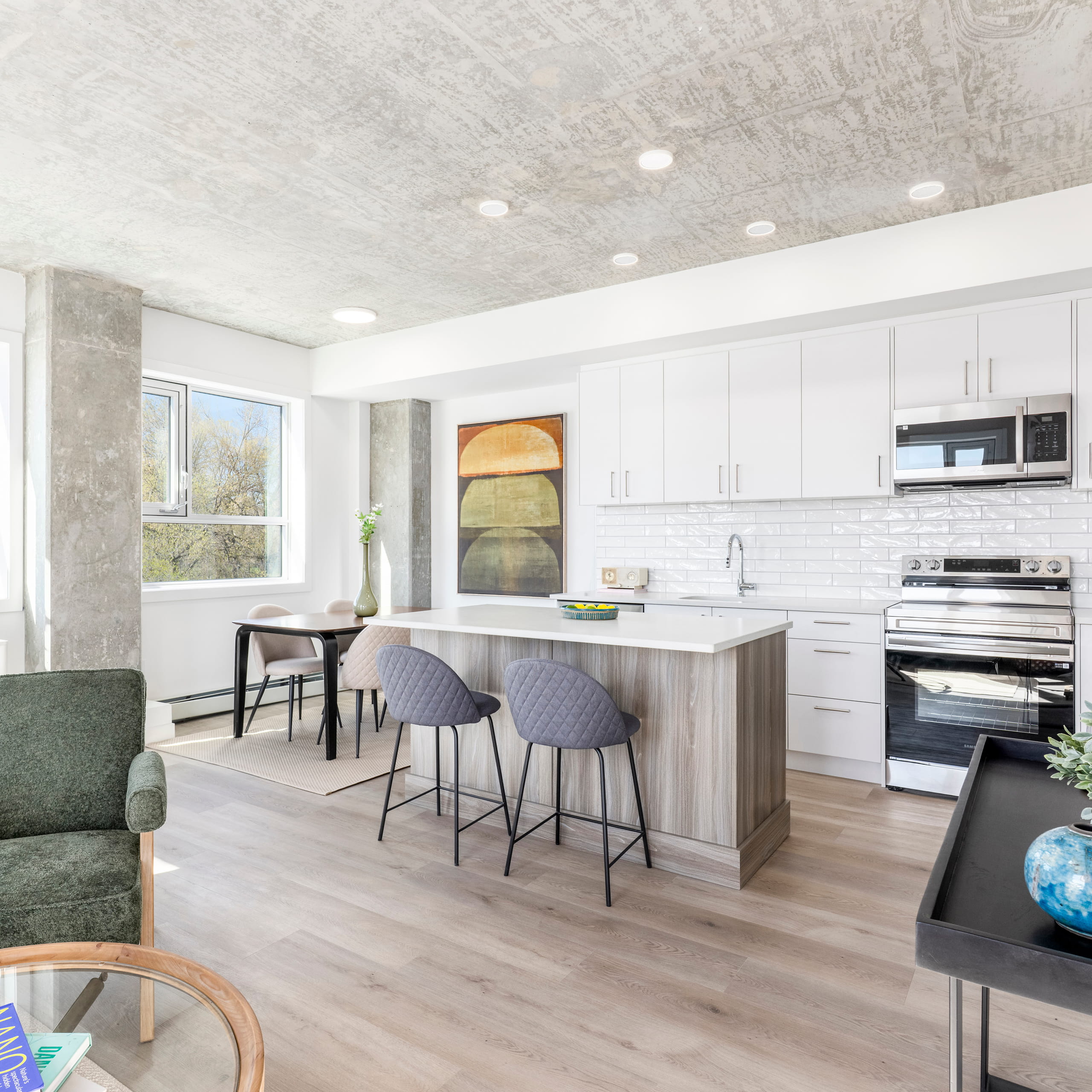Modern kitchen with white cabinets, island with two gray chairs, dining table, and concrete ceiling.