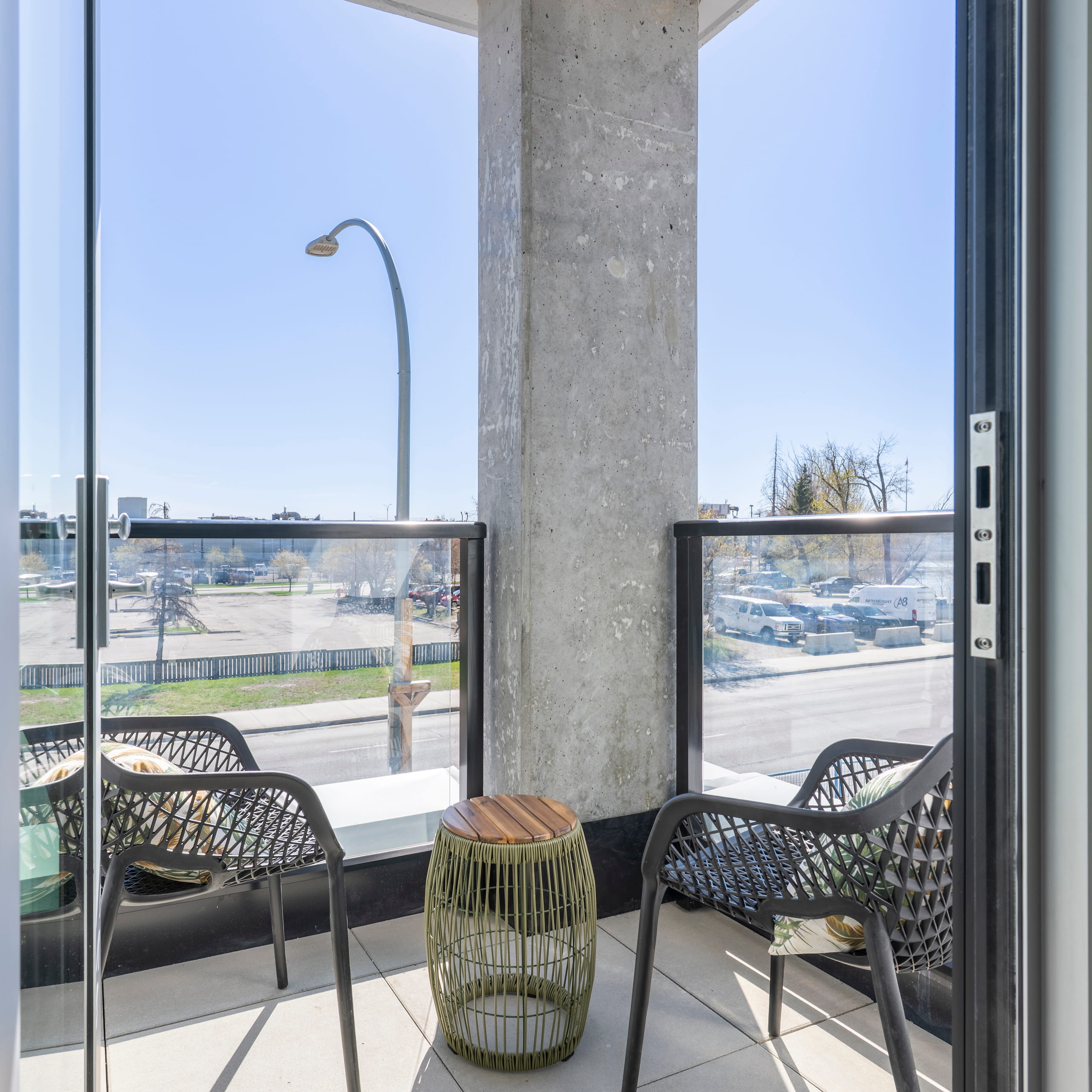 Small balcony with two black chairs, a wireframe side table, and a city street view beyond glass panels.