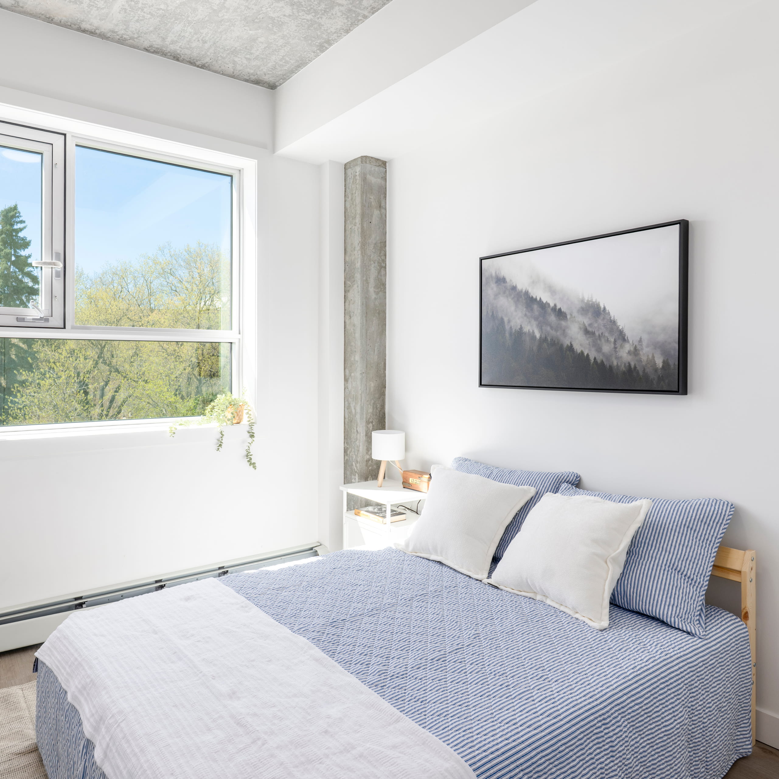 Bright bedroom with a bed in blue-striped bedding, white pillows, and forest art on the wall.