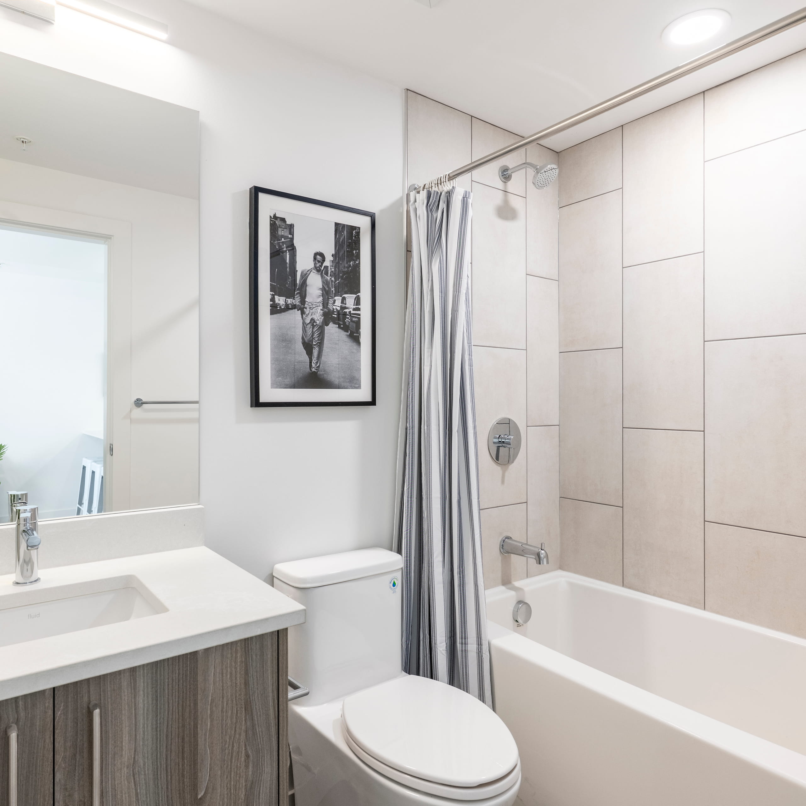 Modern bathroom with a white toilet, striped shower curtain, tiled bathtub, and black-and-white framed photo.