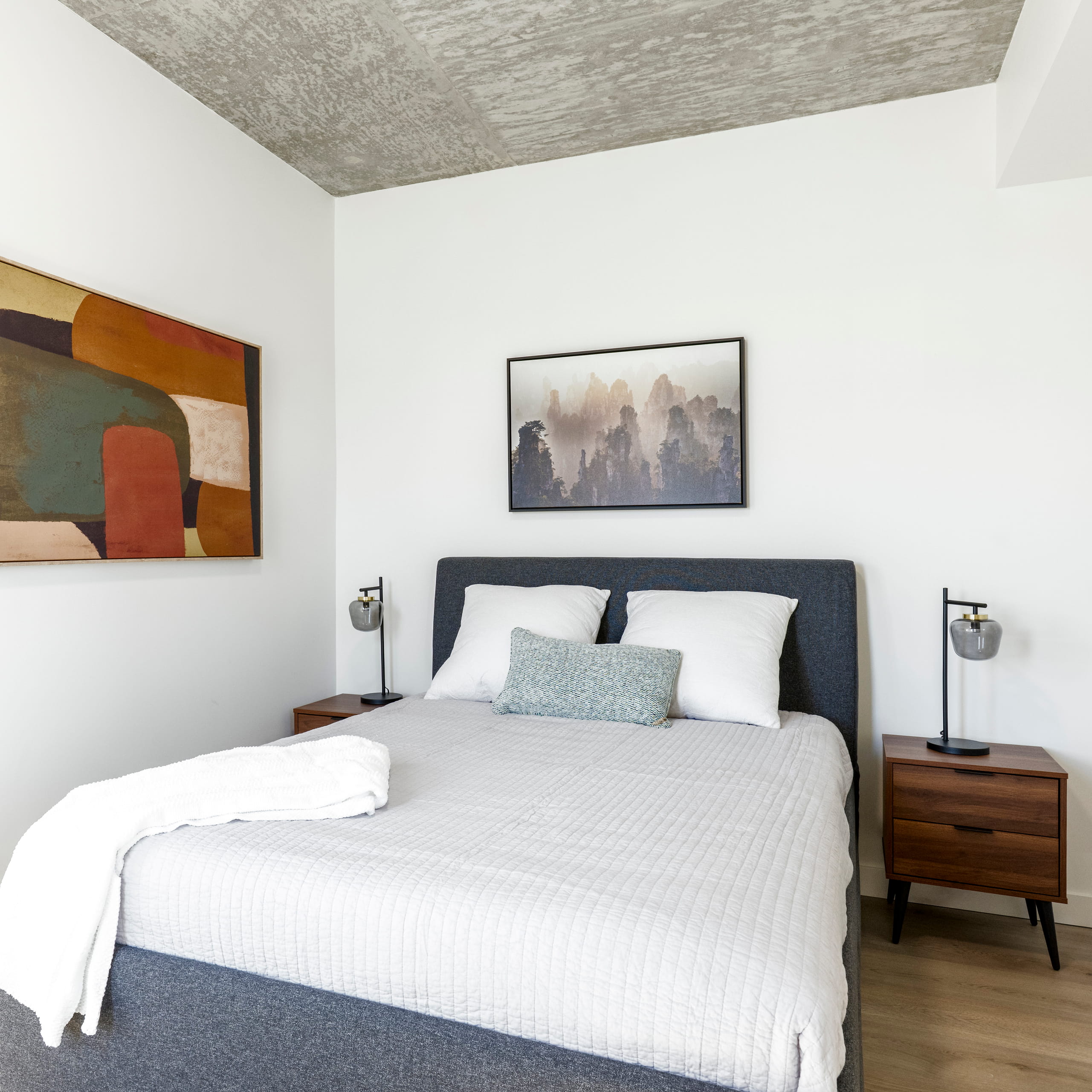 Modern bedroom with gray upholstered bed, white bedding, wood nightstands, lamps, and abstract art on walls.