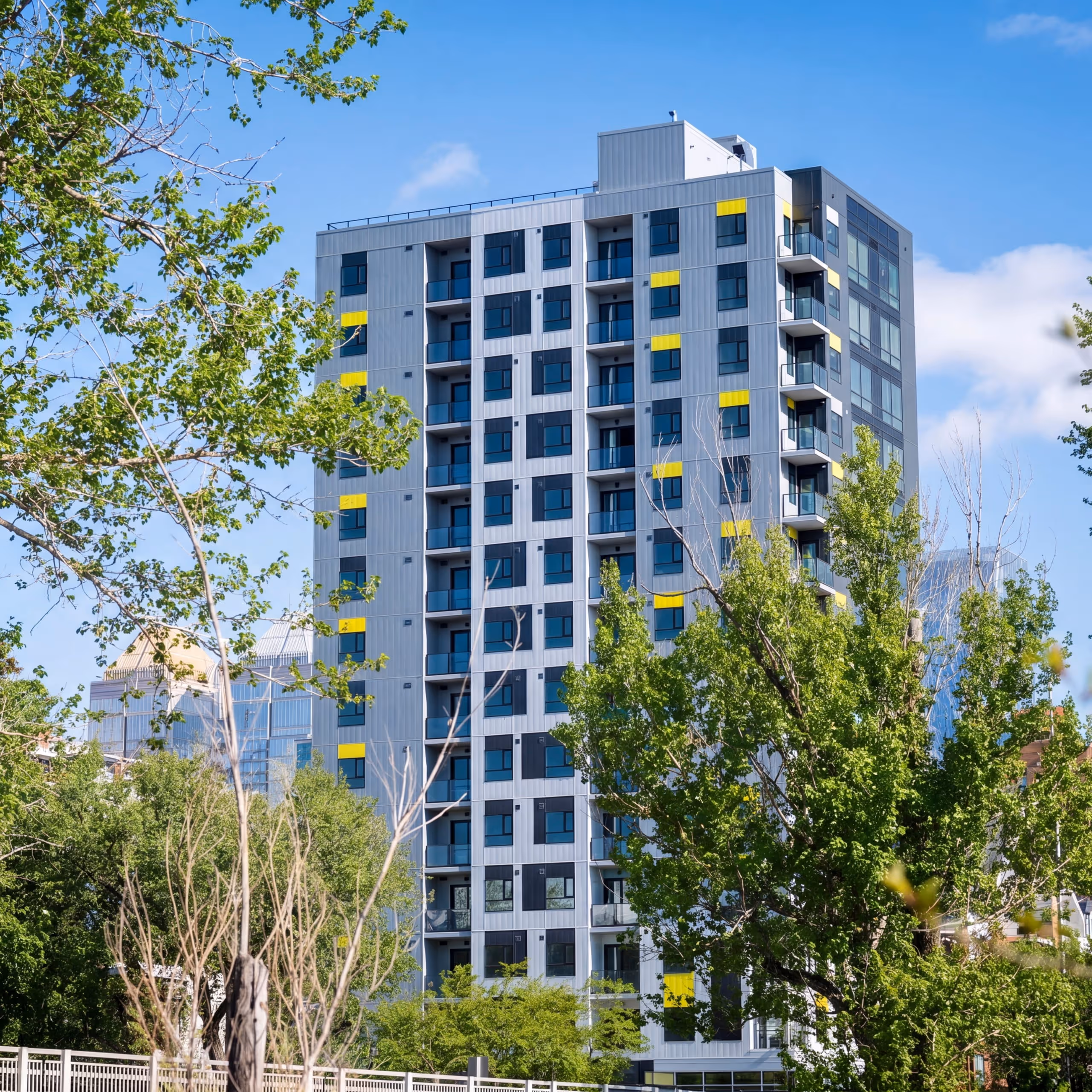 Modern tall residential building with gray facade and yellow accents surrounded by green trees under blue sky.