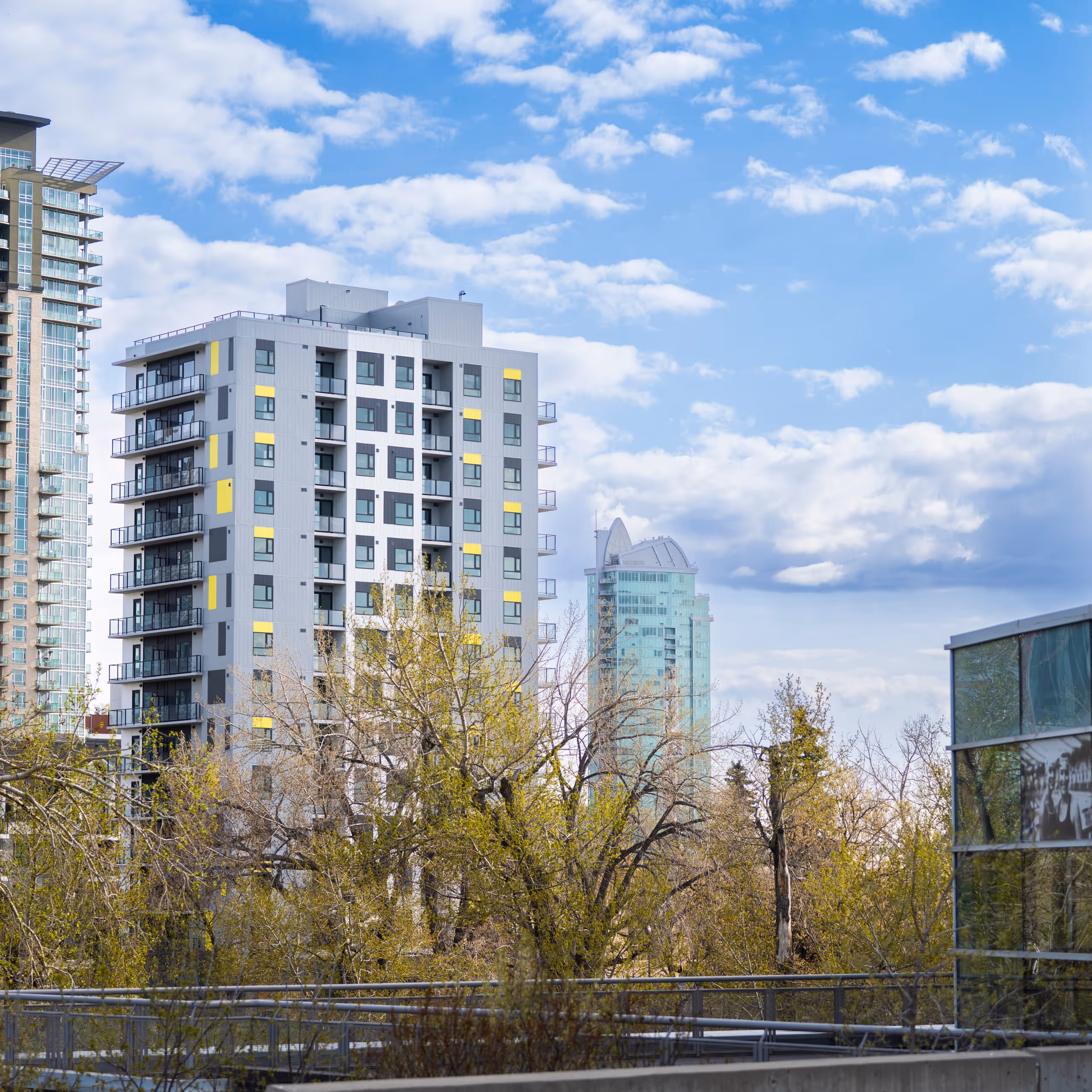 Modern apartment buildings with balconies behind leafing trees under a partly cloudy blue sky.