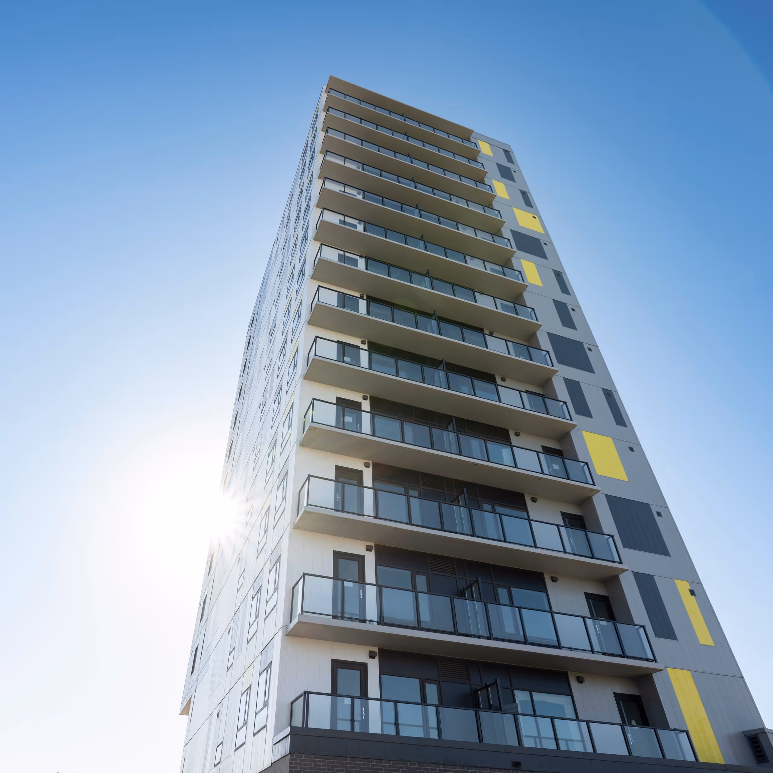 Modern high-rise building with glass balconies and yellow accents against a clear blue sky.