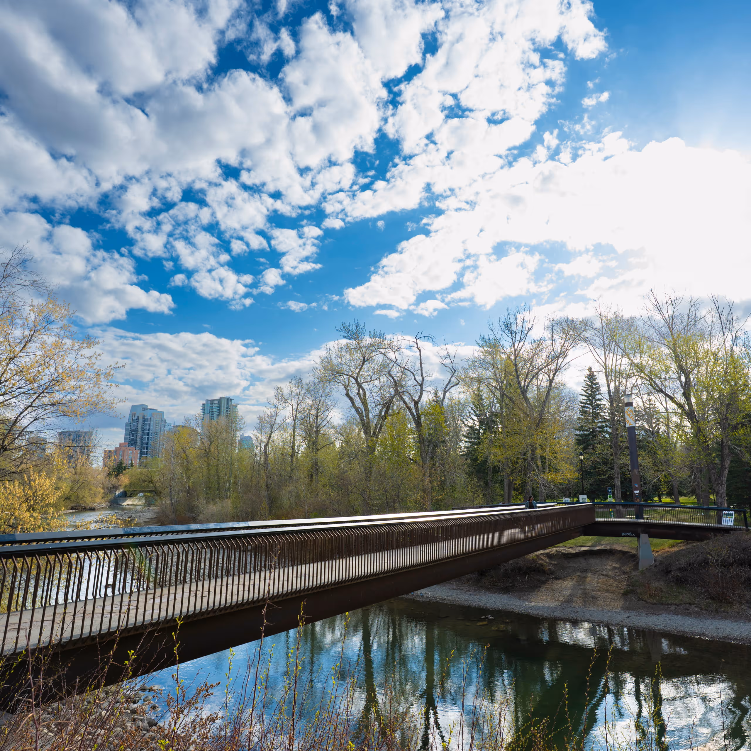Bridge crossing a calm river with trees and city buildings under a partly cloudy sky.