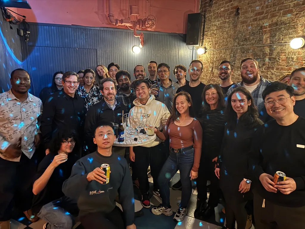 Group of people posing for a photo at a casual indoor gathering with drinks, colorful light spots, and a brick wall background.