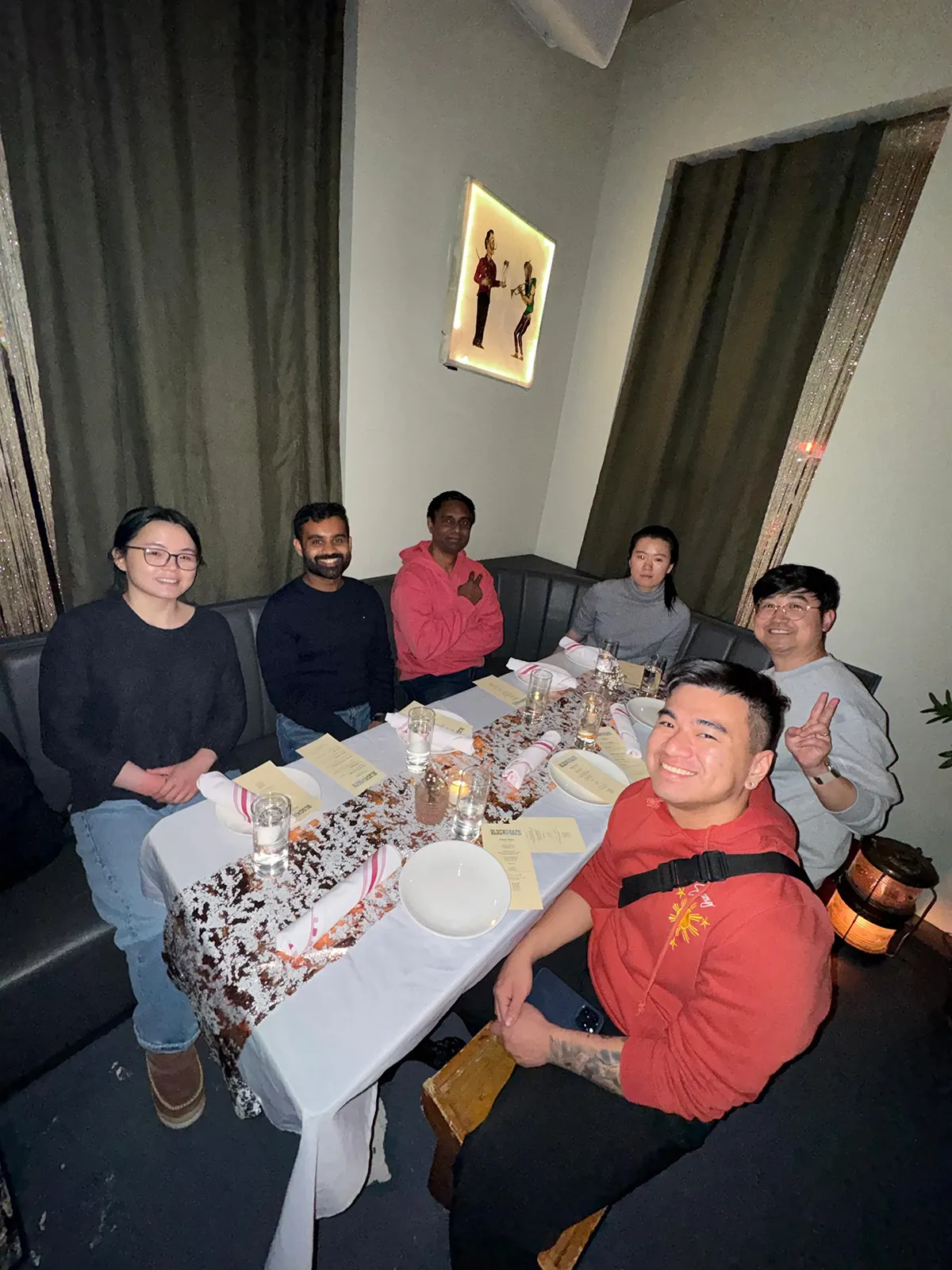 Six people seated around a decorated dining table with menus and glasses, smiling and posing for the photo in a cozy restaurant setting.