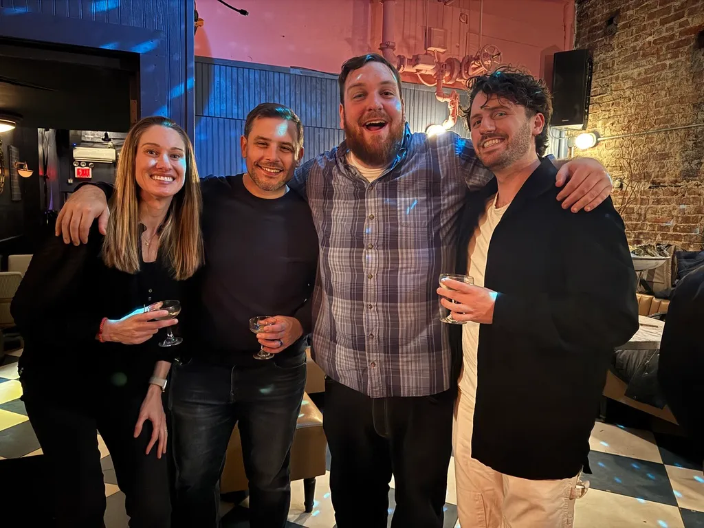 Group of four adults smiling and holding drinks in a warmly lit bar with exposed brick walls.