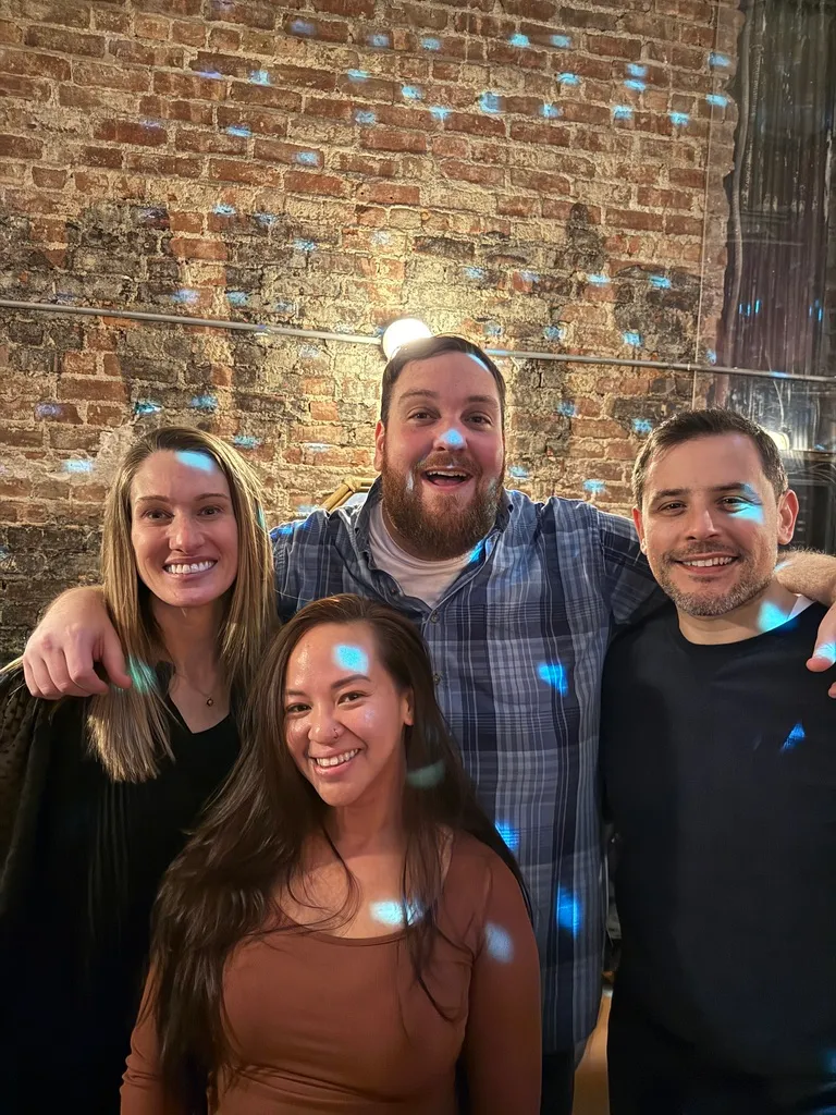 Four smiling friends standing close together indoors against a brick wall with blue light spots on them.