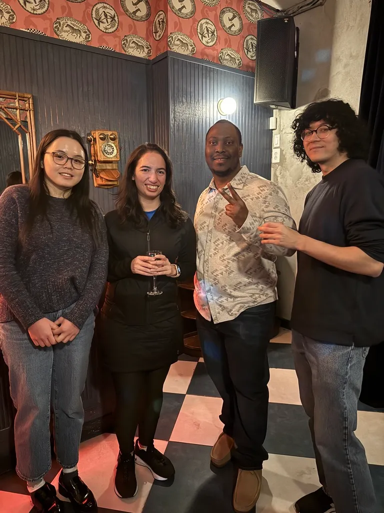 Group of four diverse people smiling indoors, two holding drinks, with patterned wallpaper and checkered floor.
