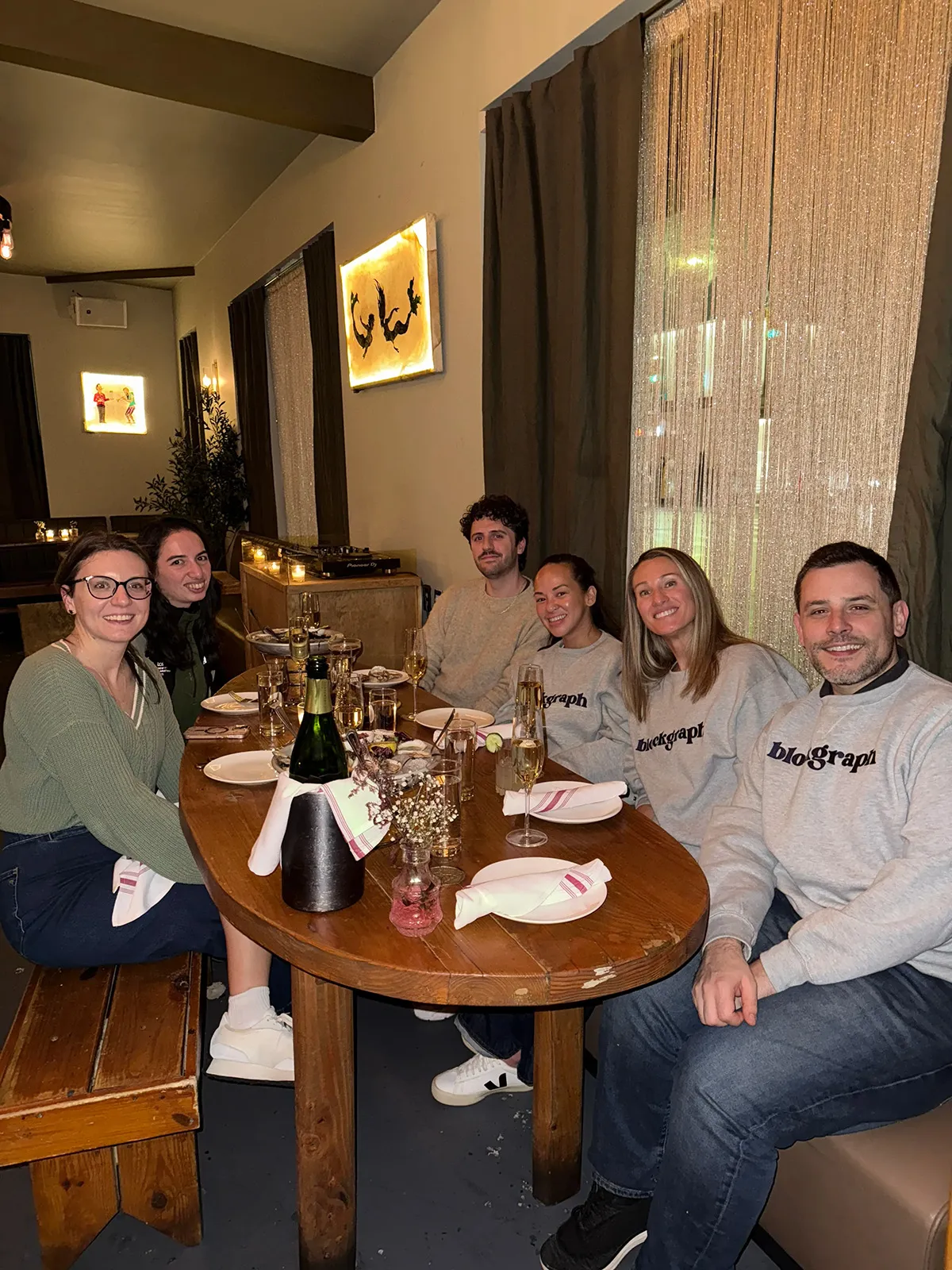 Six people sitting around a wooden dining table smiling, with drinks and plates set, in a warmly lit restaurant.