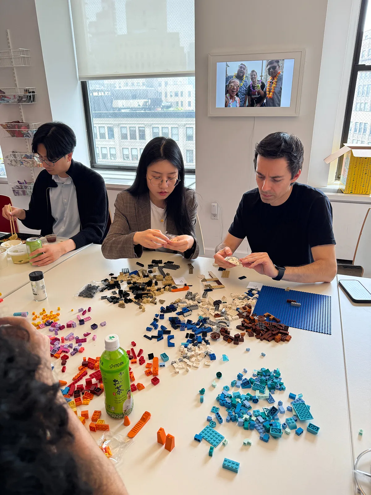 Four people sitting around a table sorting and assembling various colored LEGO bricks in a well-lit room with large windows and a framed photo on the wall.