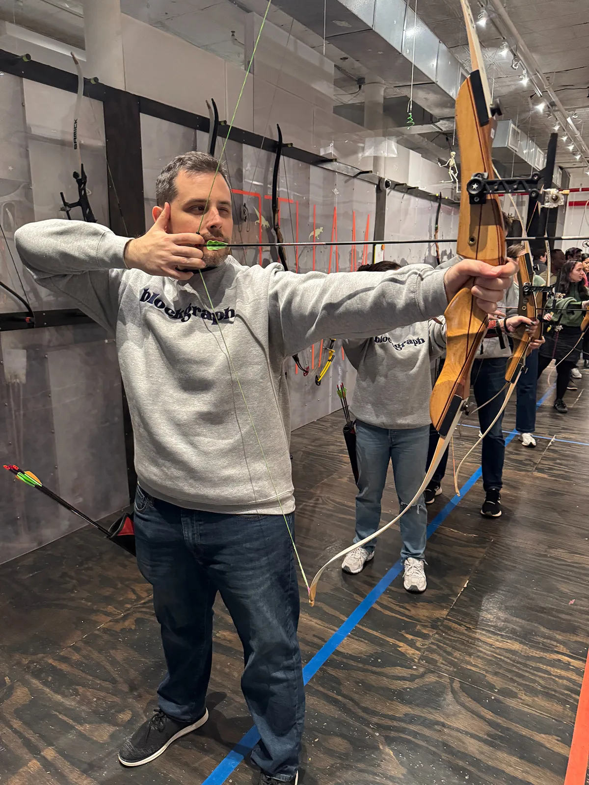 Man in grey sweatshirt and jeans aiming a wooden recurve bow in an indoor archery range.