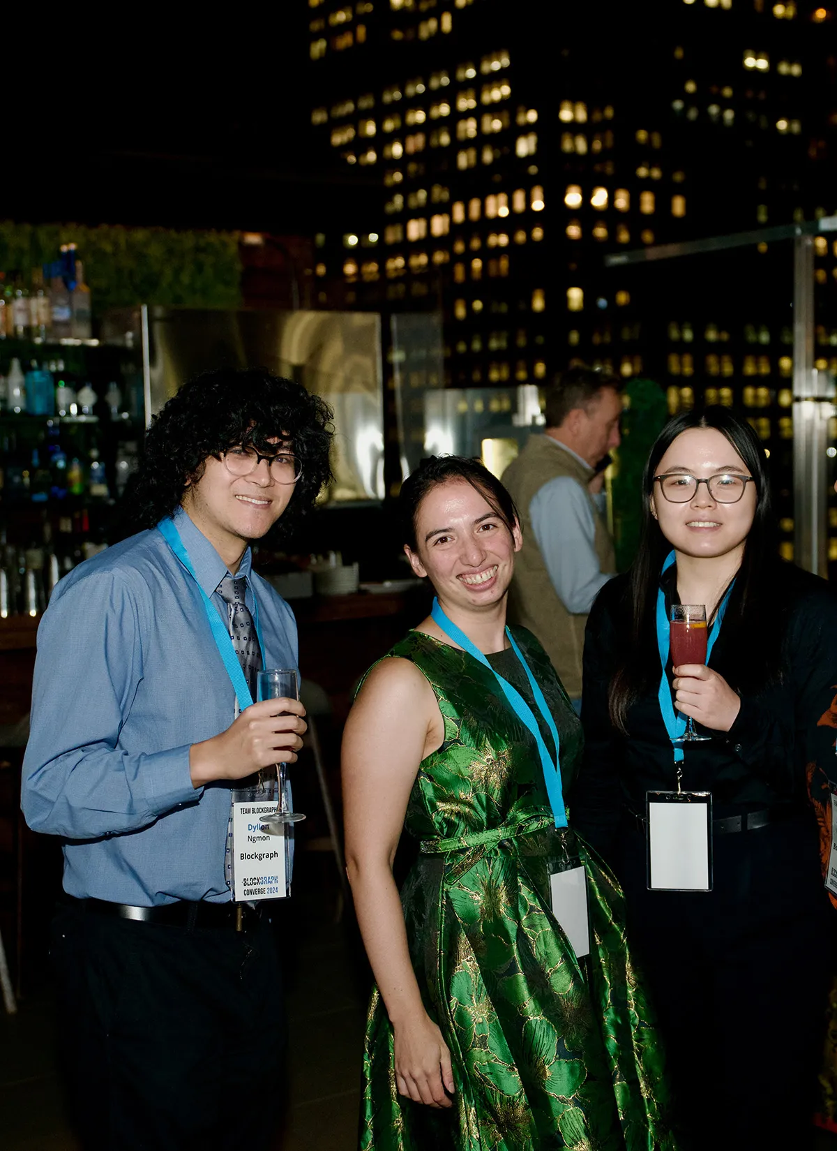 Three people wearing conference badges and lanyards smiling and holding drinks, standing inside a building at night with city lights visible through the windows.