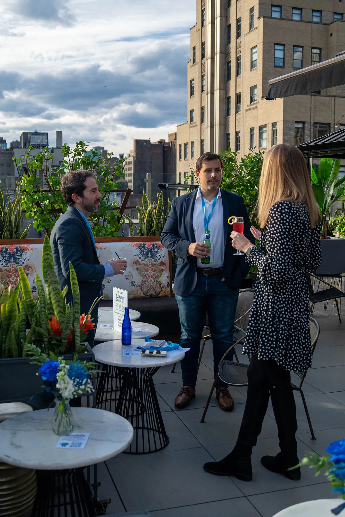 Three people having drinks and conversation on a rooftop terrace with city buildings in the background.