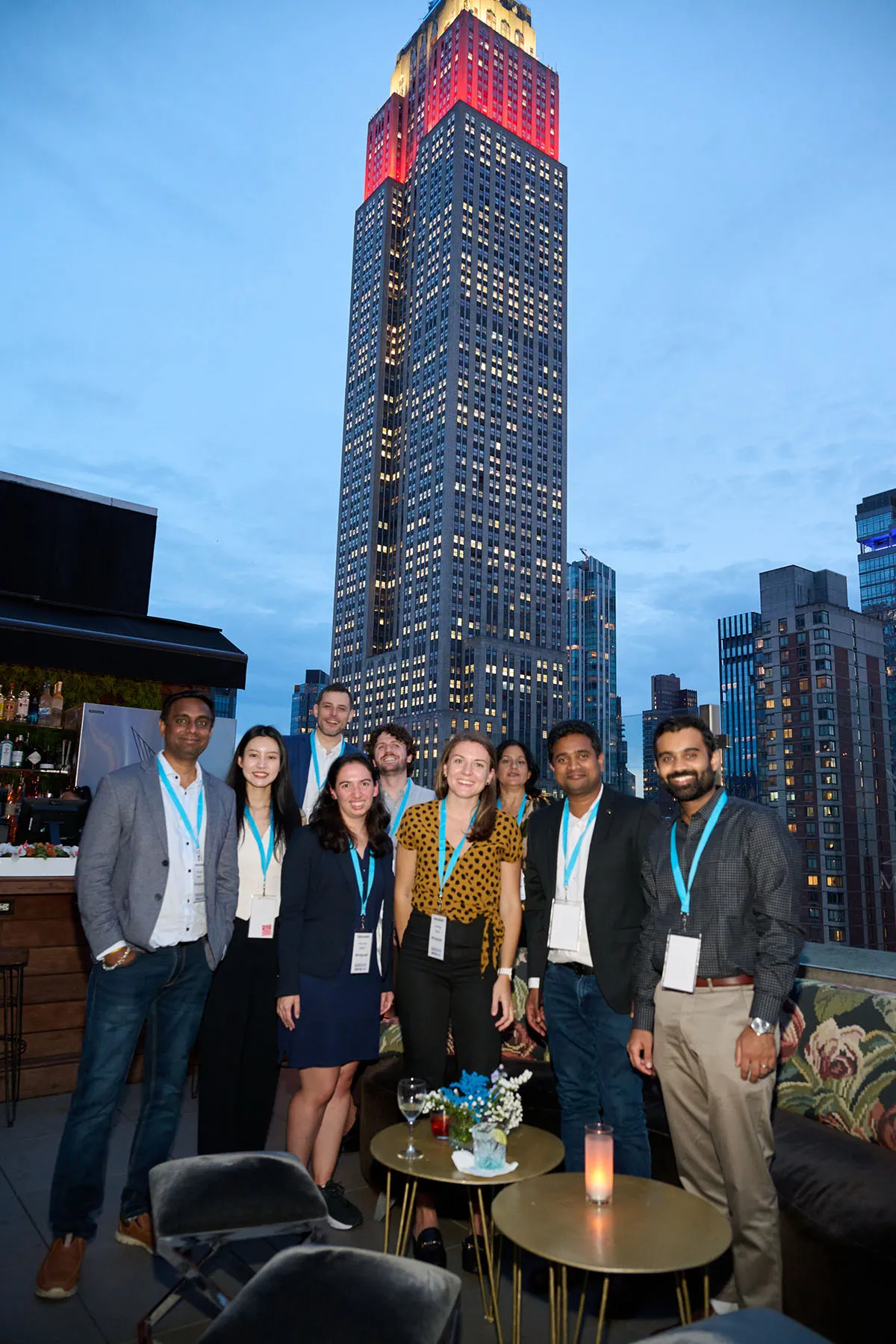 Group of nine people with conference badges posing on a rooftop with the Empire State Building lit in red and yellow in the background at dusk.