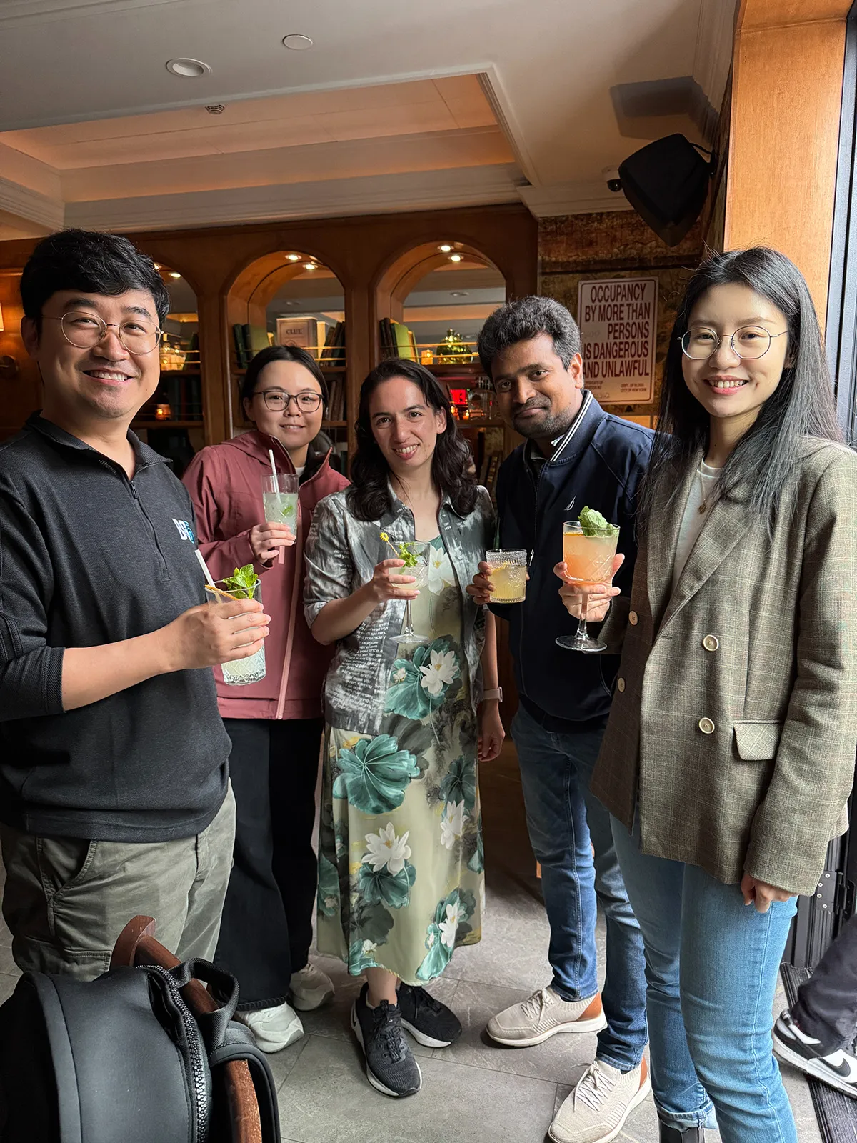 Group of five diverse people standing indoors, holding cocktails and smiling at the camera.
