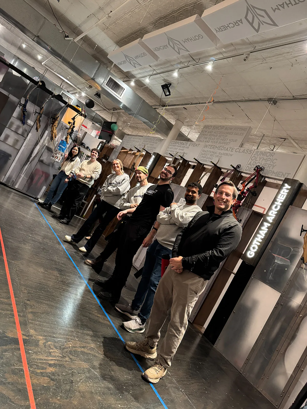 Seven people standing in a line inside an archery range with bows displayed on the wall and a Gotham Archery sign.