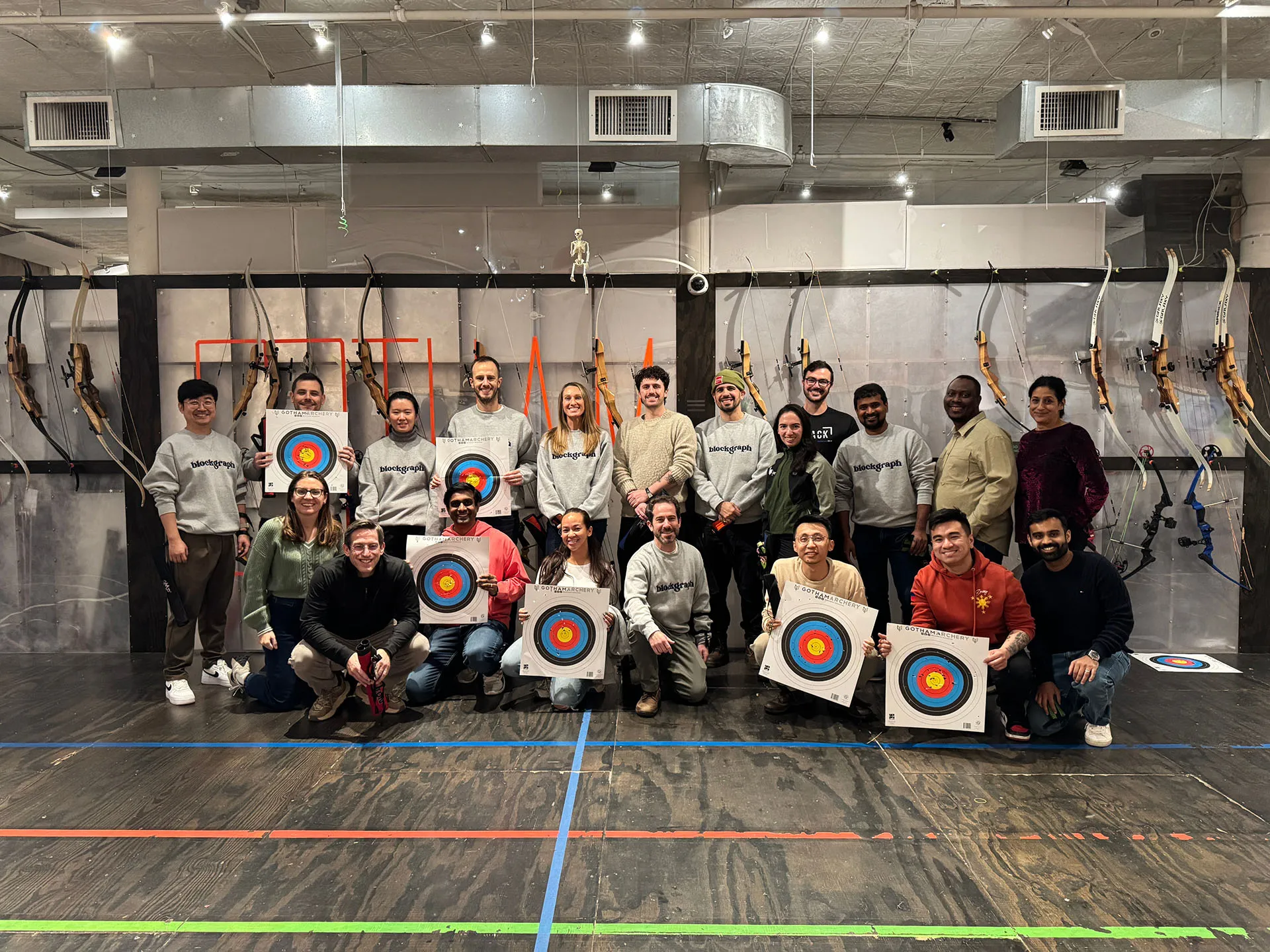 Group of people posing with archery bows and target boards in an indoor archery range.