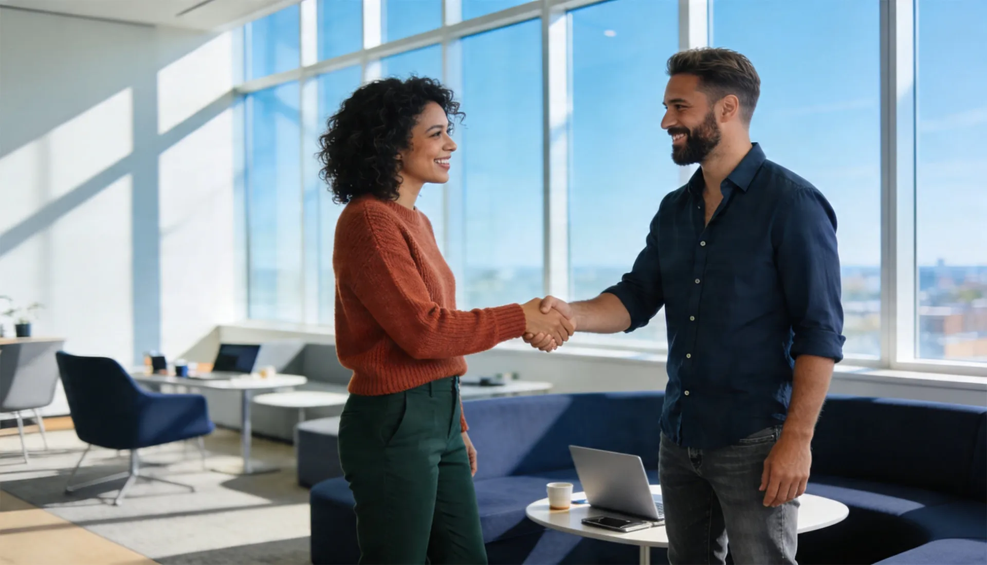 Two smiling coworkers shaking hands in a modern office with large windows and a laptop on a table nearby.