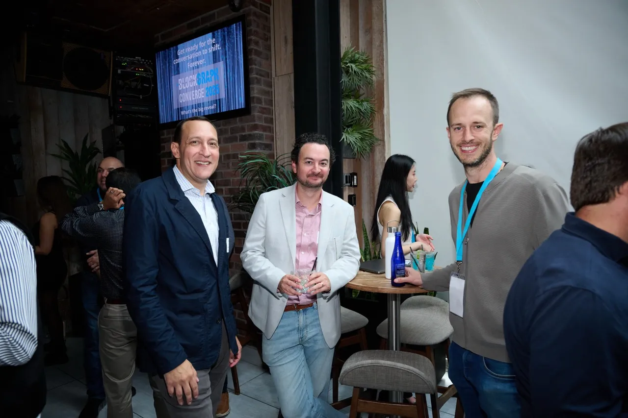 Three men smiling at a social event, with two holding drinks, standing near a high table with drinks.