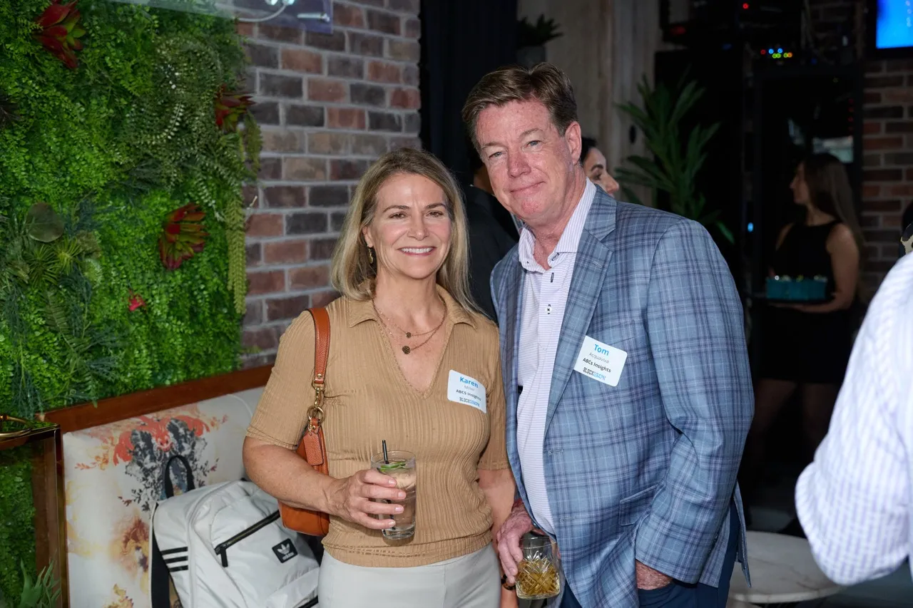 Smiling woman and man wearing name tags and holding drinks at a social event in a space with green plants and brick walls.
