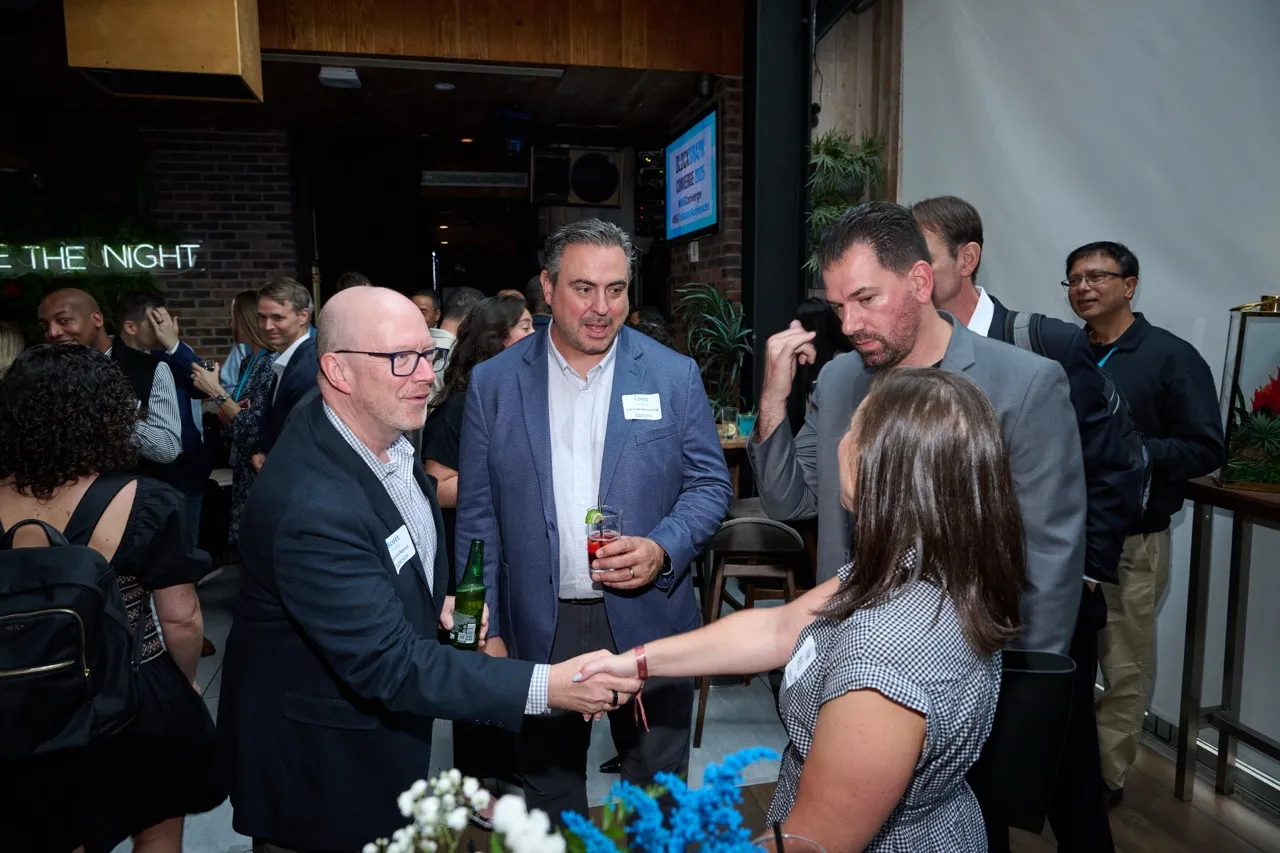 People in business attire socializing indoors, with two individuals shaking hands in the foreground.