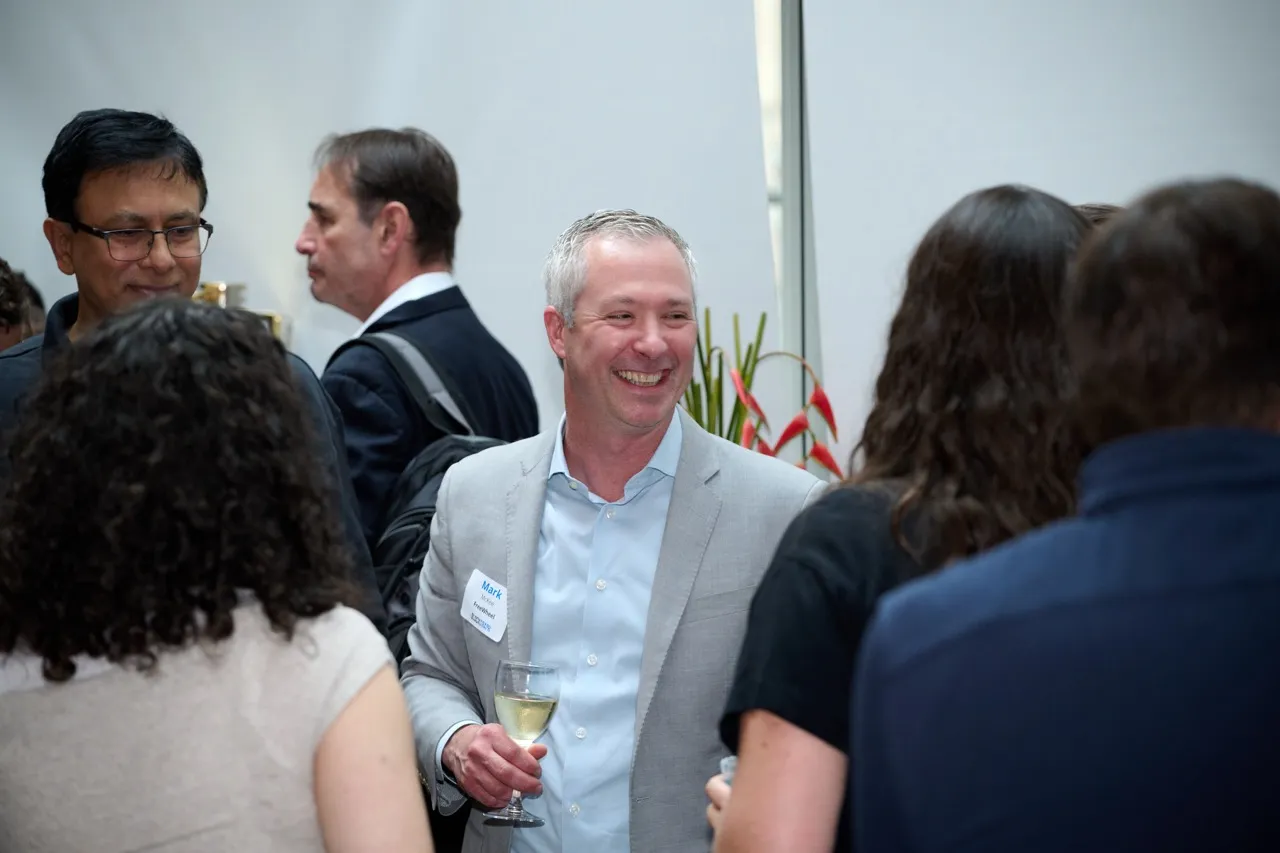Smiling man in a gray suit holding a glass of white wine, conversing with a group of people at a social event.