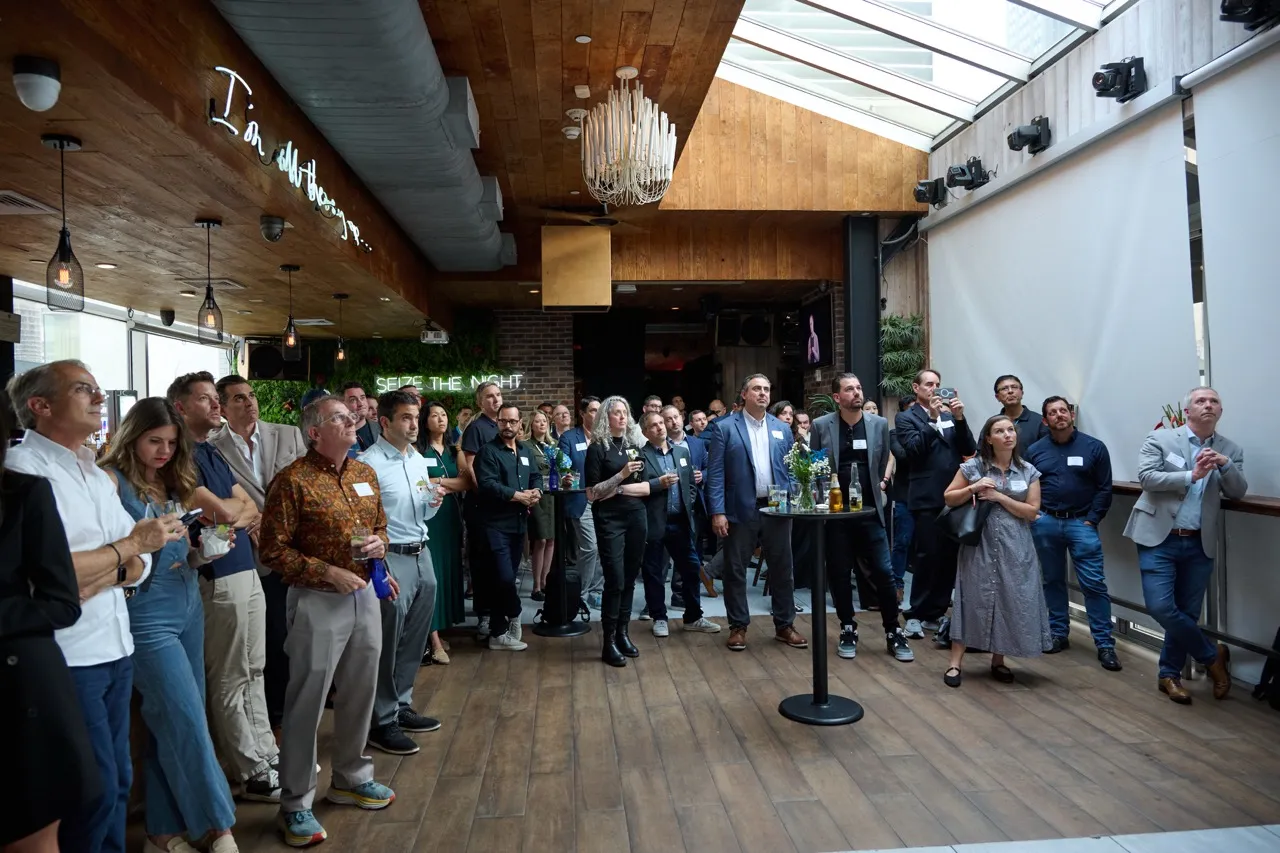 Group of diverse people standing and listening attentively in a modern indoor event space with wooden ceiling and floor, some holding drinks.