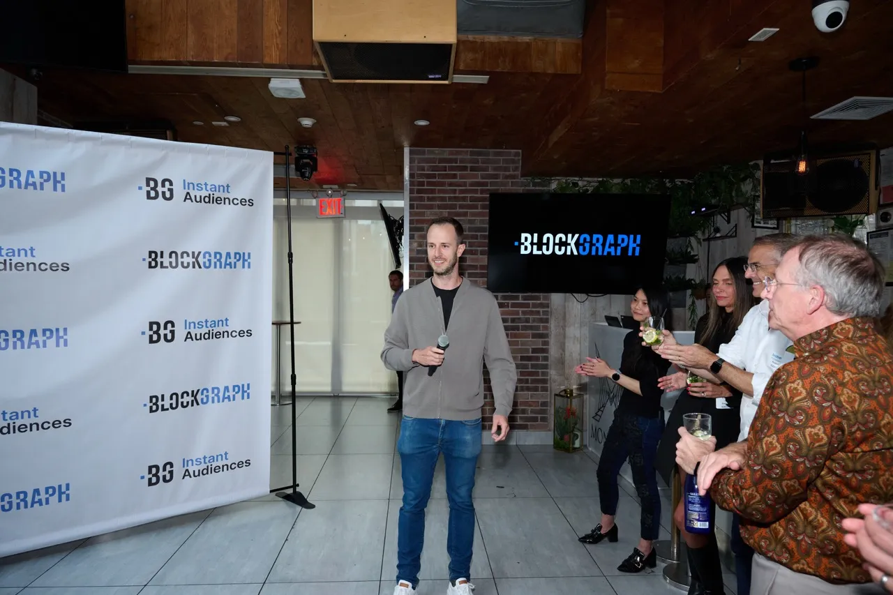 Man holding a microphone standing in front of a Blockgraph banner and screen, with a group of people holding drinks applauding nearby.