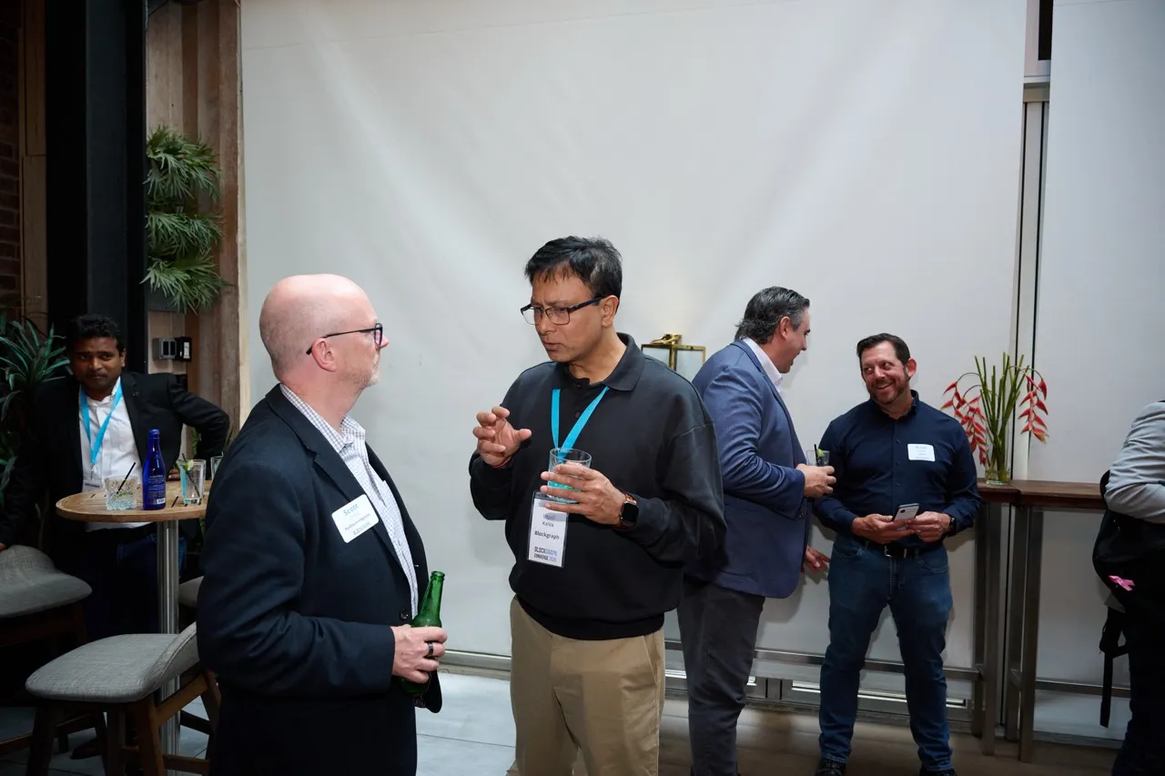 Five men engaged in conversation at a social event holding drinks, with name tags visible on two individuals.