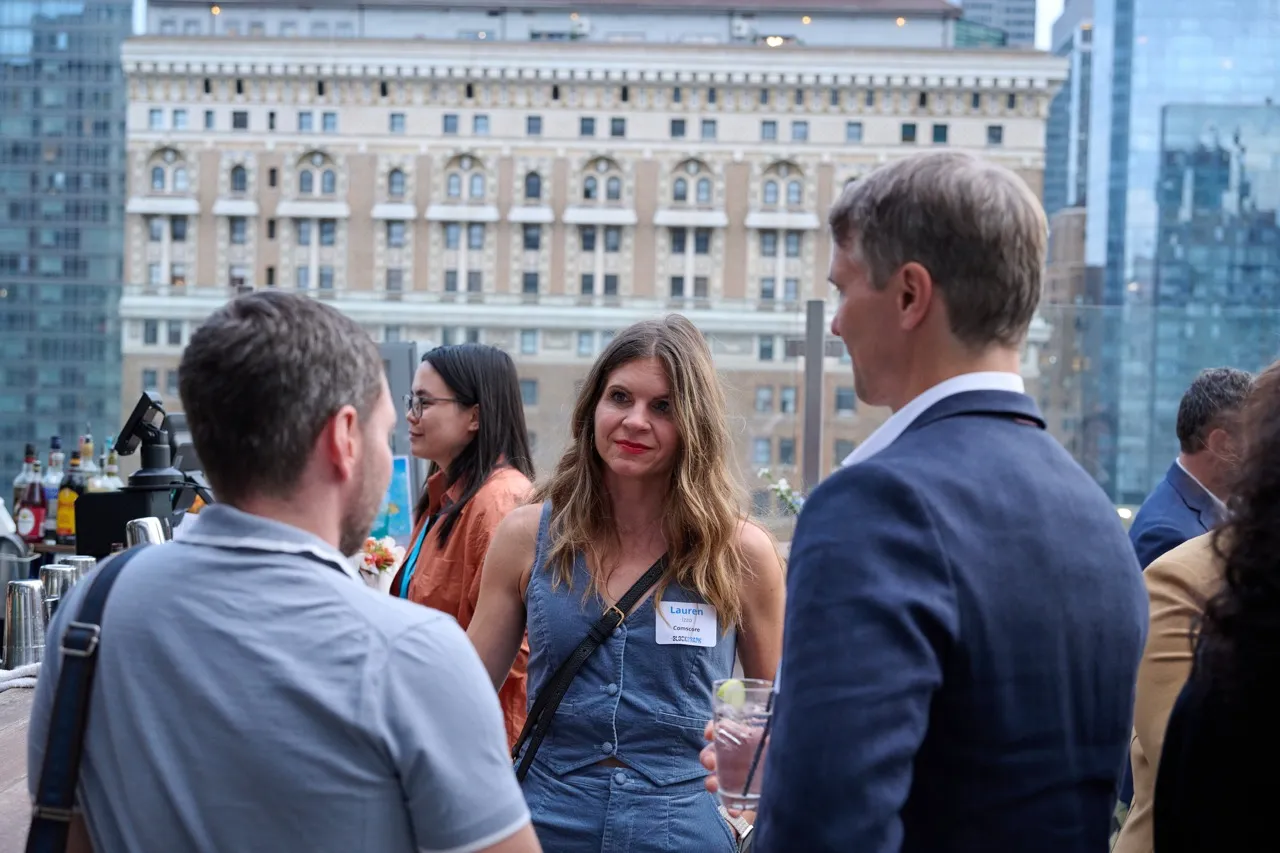 Three people conversing at an outdoor social event with a cityscape background, including a woman with a name tag reading 'Lauren.'