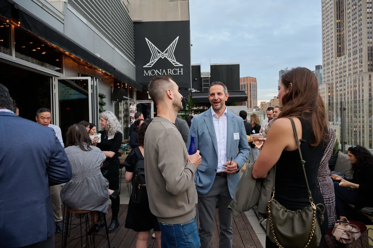 Group of people socializing and drinking on a rooftop terrace with city buildings in the background, under a Monarch sign.