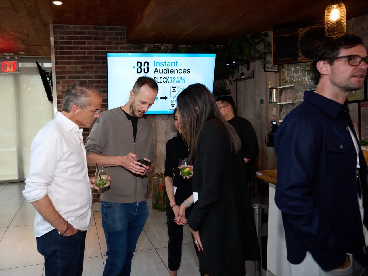 Group of five people socializing indoors, two holding drinks, with a screen in the background displaying 'BG Instant Audiences BLOCKGRAPH'.