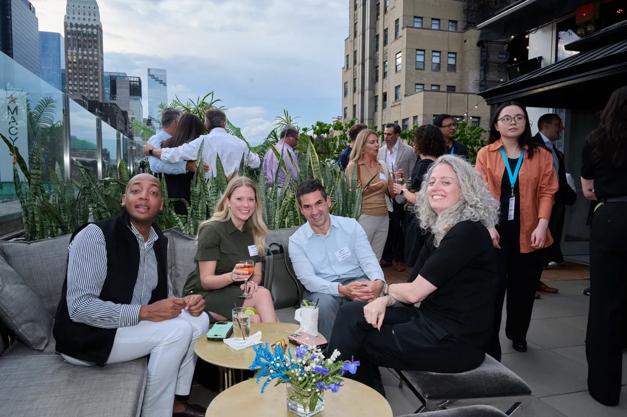 A group of four people sitting and smiling at a rooftop social event with city buildings and plants in the background.