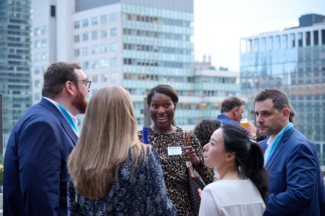 Group of professionally dressed people engaging in conversation on an outdoor terrace with city buildings in the background.