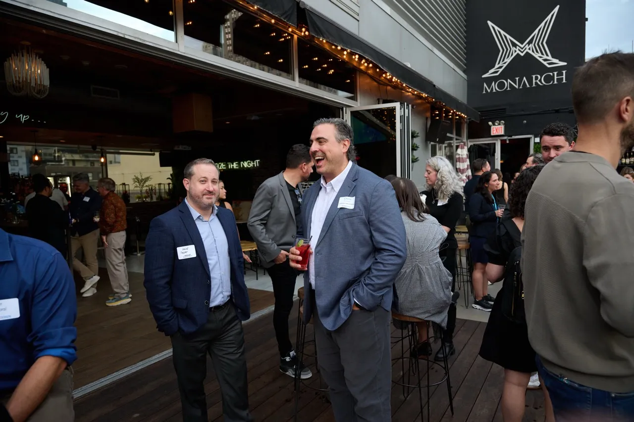 People socializing at an outdoor deck of Monarch bar, with one man holding a red drink and others chatting nearby.