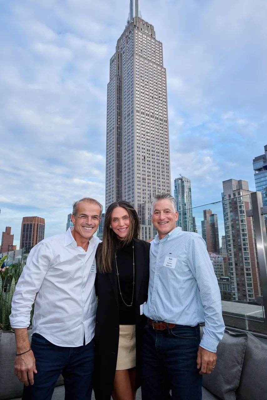 Three smiling adults posing together outdoors with the Empire State Building in the background under a partly cloudy sky.