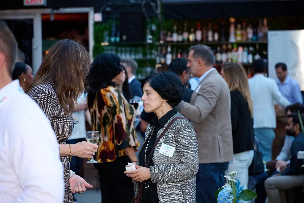 People socializing at a bar or gathering, with one woman holding a glass of white wine and another wearing a name tag.