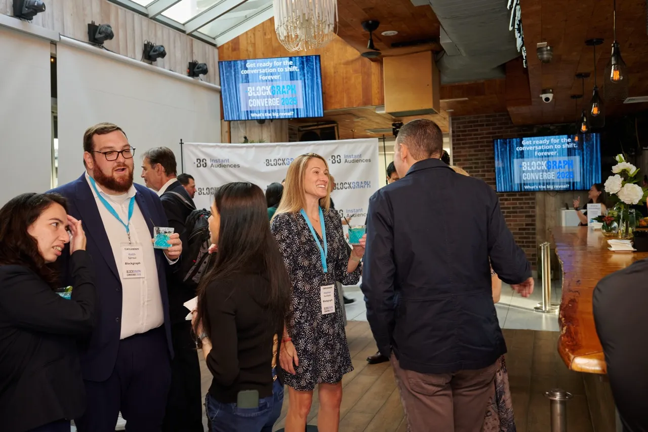 People networking and conversing at a Blockgraph Converge 2023 event in a modern, well-lit venue with screens displaying event information.