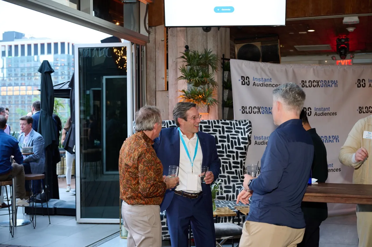 Three men standing and conversing at an indoor-outdoor event space with a Blockgraph banner in the background.