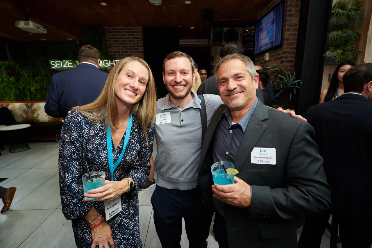 Three smiling adults at a social event holding blue cocktails, two wearing name tags labeled Jesse and Joe.