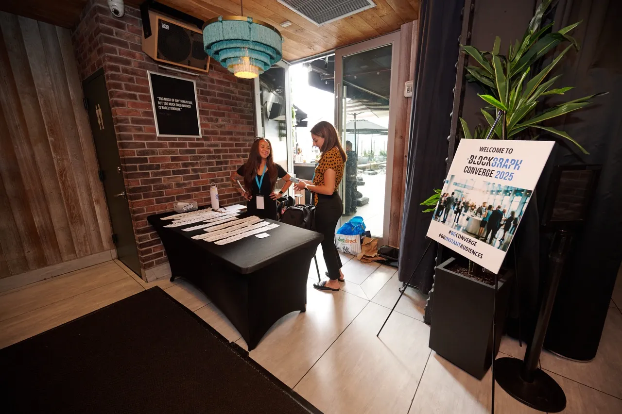 Two women standing behind a table covered with name tags at a registration area for Blockgraph Converge 2025 event, with a welcome sign nearby and a brick wall background.