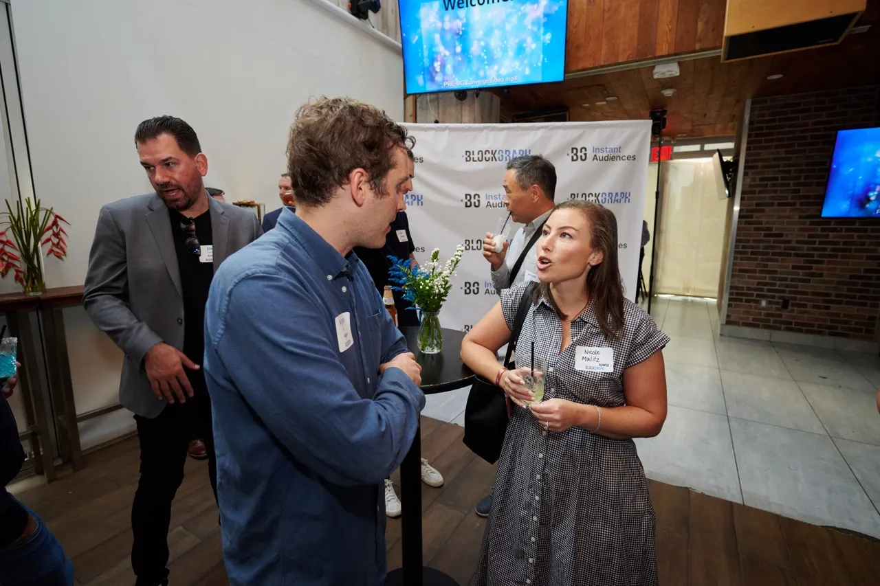 Group of people socializing indoors at a professional event with name tags and drinks near a table with flowers and a Blockgraph banner.