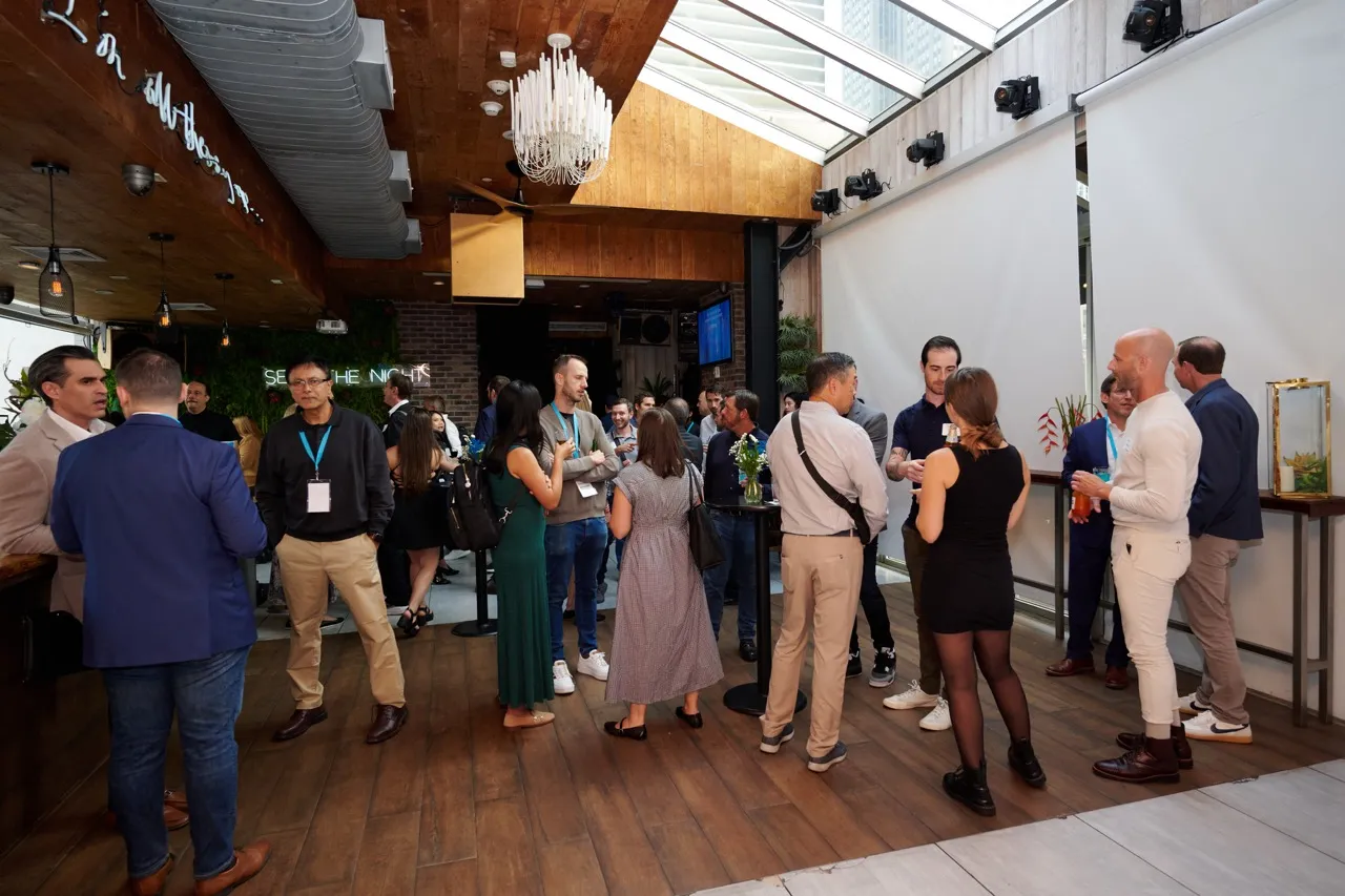 Group of adults socializing in a modern indoor venue with wooden floors and ceiling, gathered around high tables with drinks.